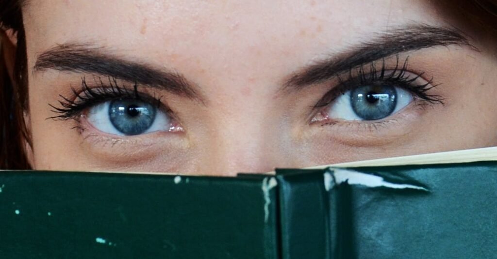 Close-up of a woman with blue eyes peeking over a book, portraying curiosity.